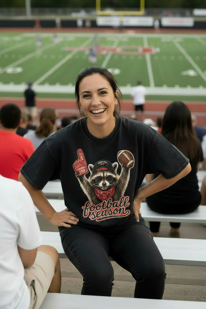 A black acid-wash graphic t-shirt featuring a raccoon holding a foam finger and a football, with 'football season' written in red script below it.